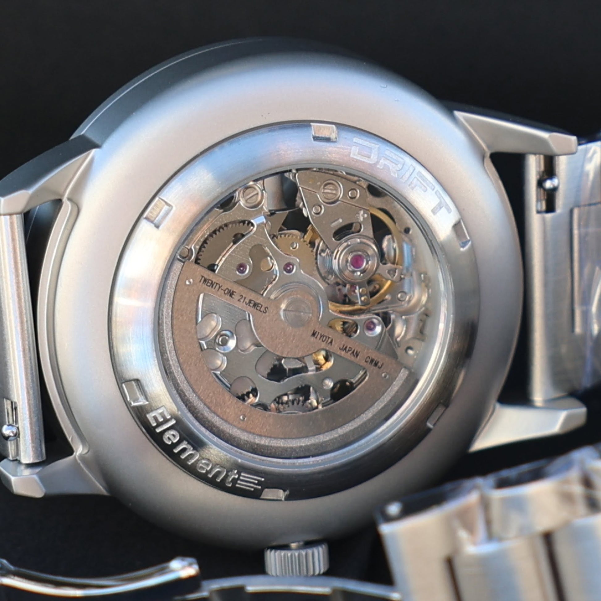 The back view of a DriftElement PGT Essen Motorshow rim watch, displaying the intricate mechanical movement and craftsmanship. 'DRIFT Element' is etched around the transparent case back, showcasing the watch's engineering that includes 'TWENTY-ONE JEWELS' and precision components. This highlights the young German startup's dedication to innovative design and quality watchmaking.