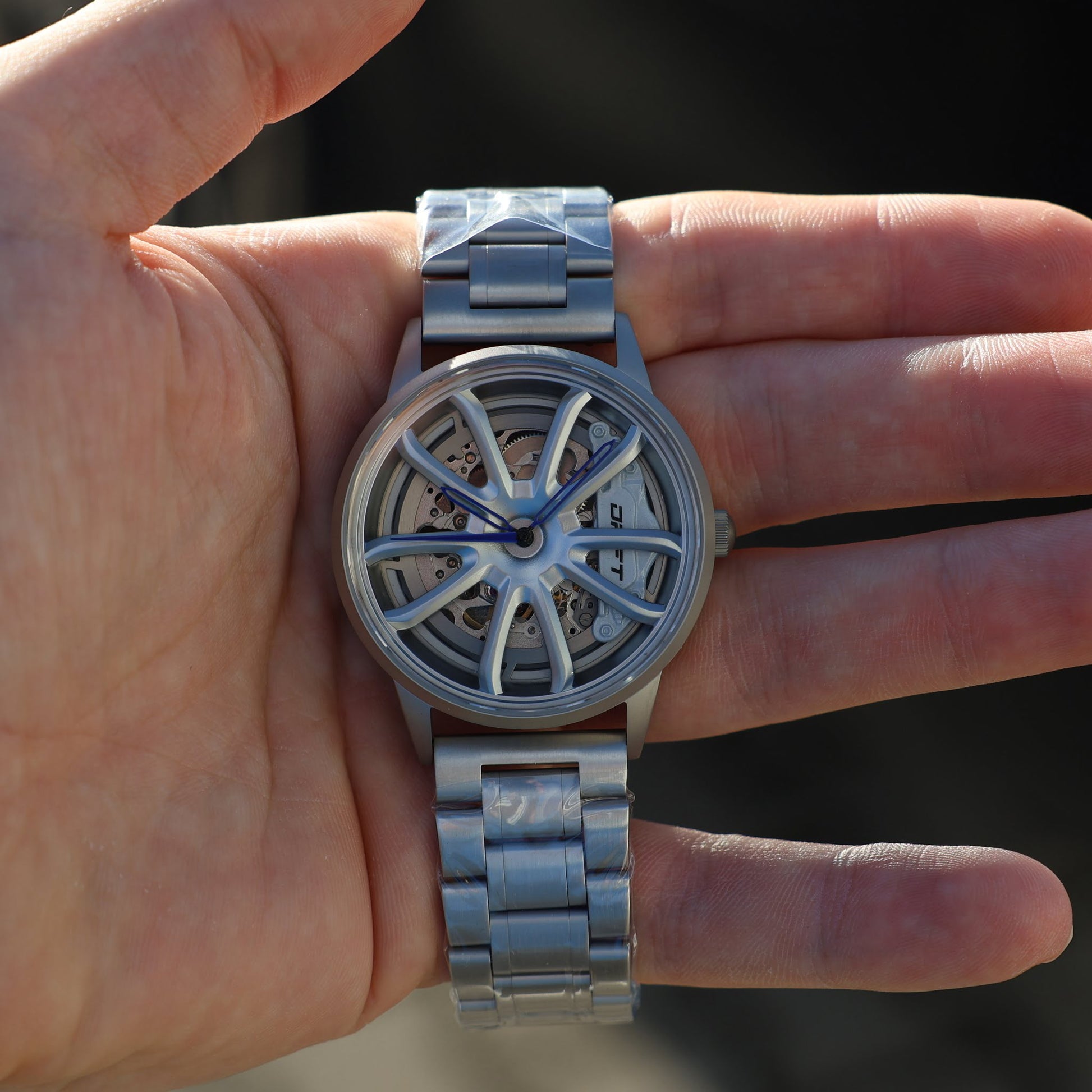 This particular model, suitable for showcasing at events like the PGT Essen Motor Show, features a distinct wheel rim-inspired face, with the watch mechanisms visible through the rim patterns. It has a silver-tone metal bracelet and a case, with blue watch hands adding a pop of color to the design. This watch is a prime example of DriftElement's commitment to combining automotive passion with horology, presenting a design that is both contemporary and mechanically driven.