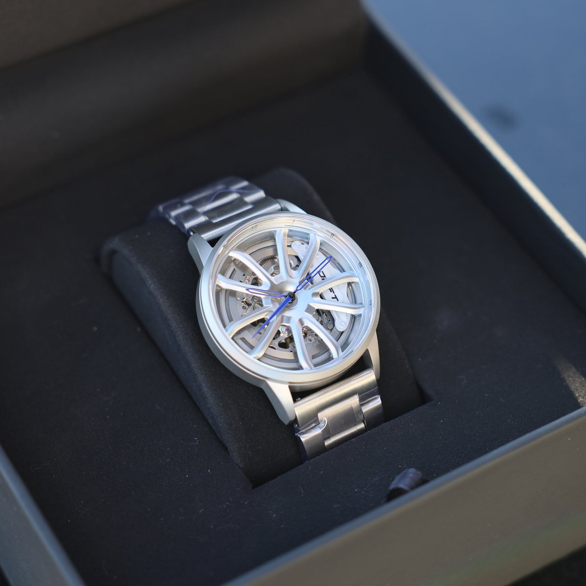 A sophisticated PGT Essen Motorshow rim watch by DriftElement, resting in an elegant black presentation box. The timepiece showcases a silver-tone bracelet and a transparent, wheel-shaped dial through which the mechanical movements are visible. Blue hands add a vibrant touch to the watch, epitomizing DriftElement's commitment to innovative design in their Germany-based startup.