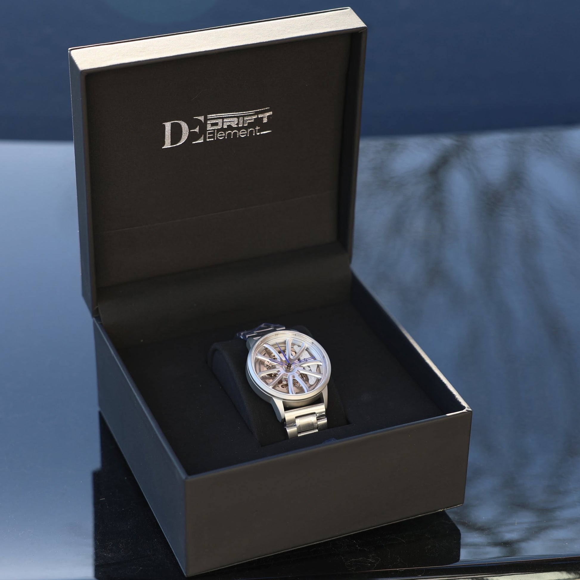An elegant PGT Essen Motorshow wheel-designed watch by DriftElement is presented inside a sophisticated black box with the company's logo embossed in silver on the inside lid. The watch itself features a metallic bracelet and a complex, wheel-inspired dial that showcases the intricate craftsmanship. It is a reflection of the German startup's commitment to innovative and cutting-edge design in watchmaking