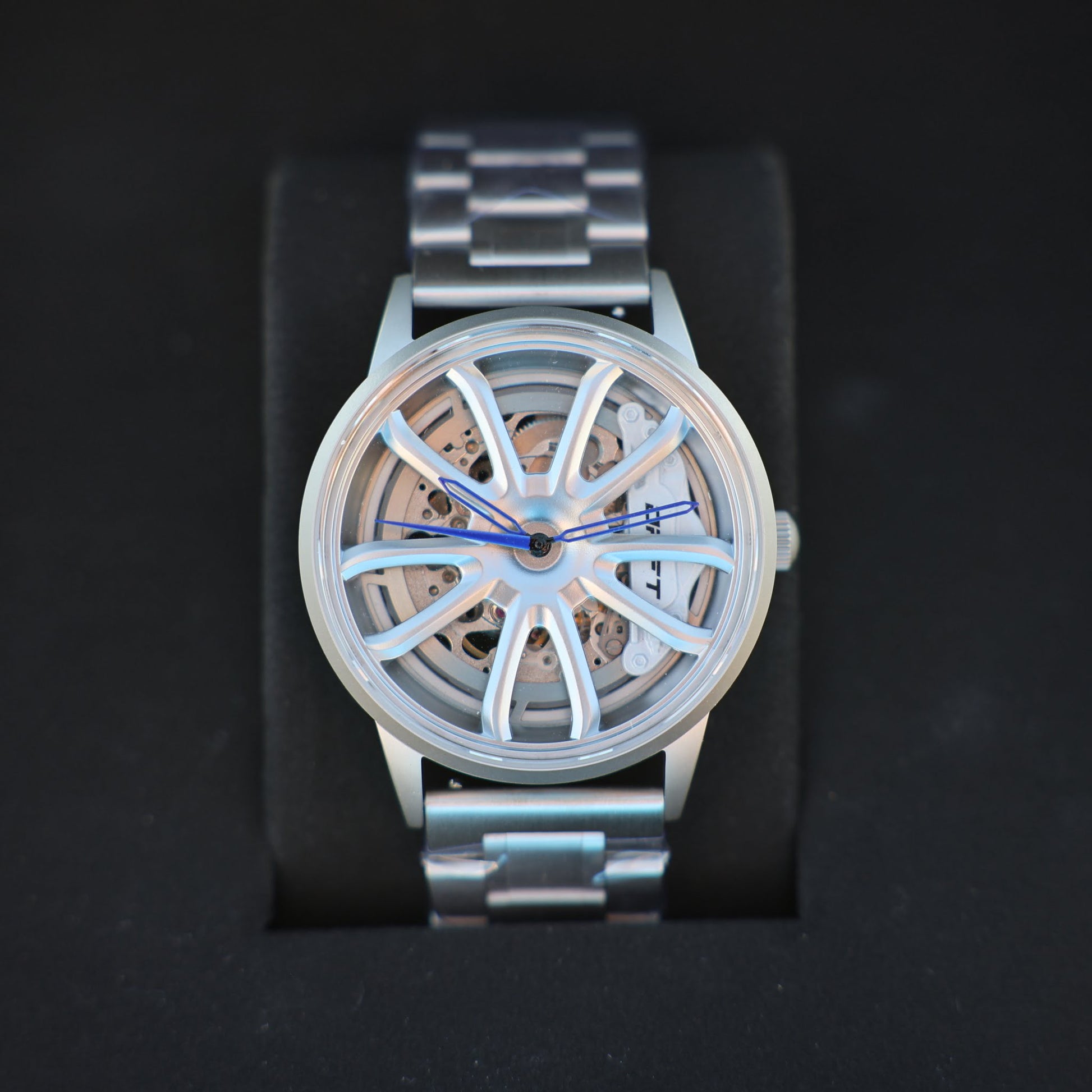 An elegant PGT Essen Motorshow wheel watch by DriftElement displayed on a black stand. This watch has a silver band and casing with a unique, open-worked dial resembling a car wheel, complete with intricate mechanical detailing visible beneath. Blue watch hands add a subtle pop of color to the sophisticated design, characteristic of the innovative and automotive-inspired timepieces from the German startup.