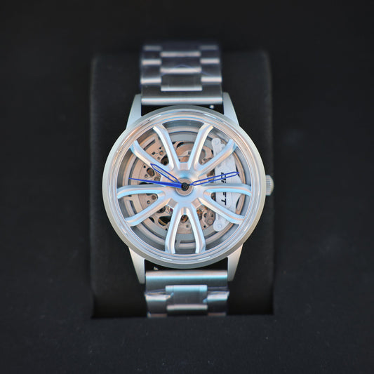 An elegant PGT Essen Motorshow wheel watch by DriftElement displayed on a black stand. This watch has a silver band and casing with a unique, open-worked dial resembling a car wheel, complete with intricate mechanical detailing visible beneath. Blue watch hands add a subtle pop of color to the sophisticated design, characteristic of the innovative and automotive-inspired timepieces from the German startup.