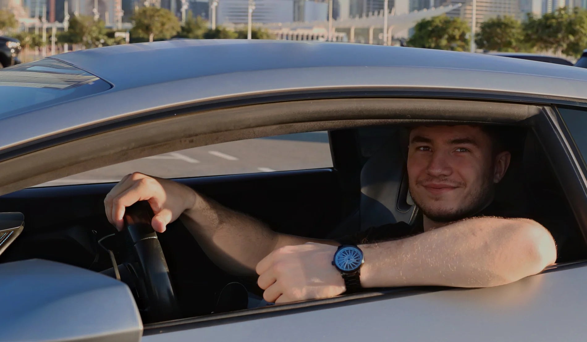 A young man smiles from the driver's seat of a car, wearing a DriftElement watch with a bold rim design on his wrist. The innovative style of the watch reflects the German startup's aim to appeal to car enthusiasts with its unique design.