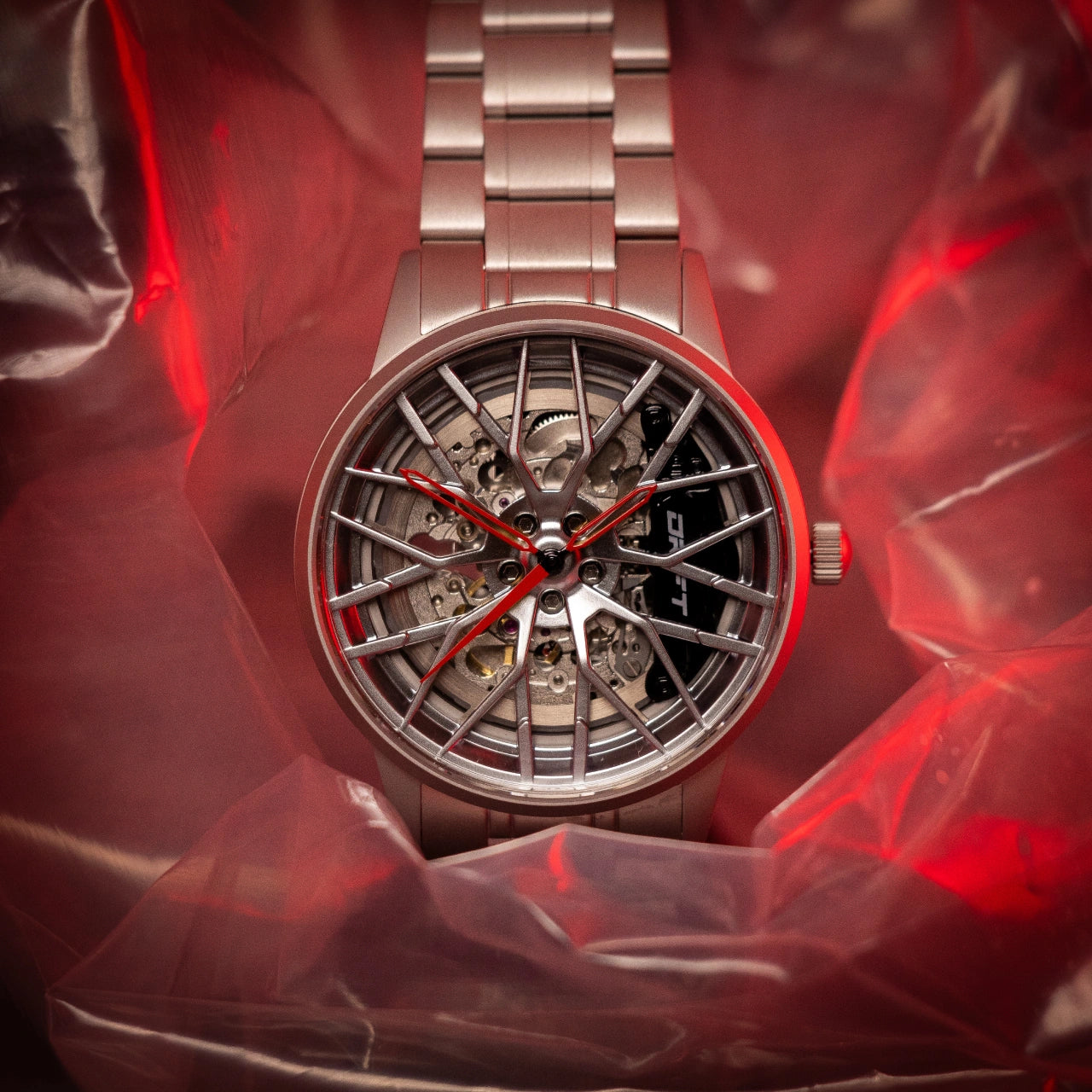 The image showcases an Automatic Motorsport EMS Edition wristwatch by DriftElement, set against a red backdrop. This timepiece features a wheel rim-inspired design, complete with visible mechanical components that highlight the intricacy of its engineering. Red hands on the watch face add a striking contrast, enhancing its automotive aesthetic. DriftElement, a fresh German startup, is the creator of this innovative watch, reflecting their dedication to unique and forward-thinking design.