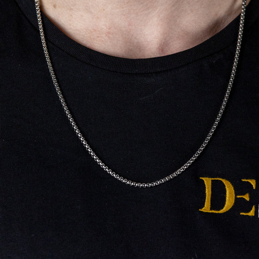 The image captures a close-up of a slender, silver chain necklace worn by someone in a black crew neck shirt with the golden lettering 'DE' visible, suggesting it may be branded apparel. The necklace's simplicity complements the implied innovative and stylish design ethos of DriftElement, a youthful German startup known for creating watches with unique rim designs, reflecting their commitment to both elegance and automotive-inspired aesthetics.