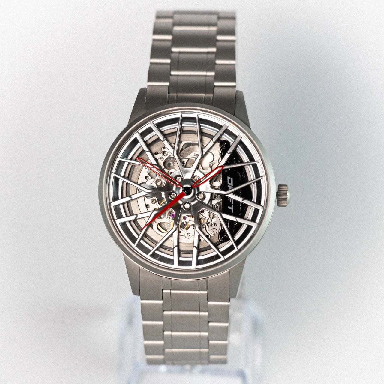 The image displays an Automatic Motorsport EMS Edition wristwatch. The watch features a unique wheel rim design, symbolizing DriftElement's innovative approach to timepieces. The metallic structure, visible gears, and the striking contrast of silver and red accents exemplify the creative fusion of horology and automotive design. DriftElement is a German startup known for such distinctive watches.