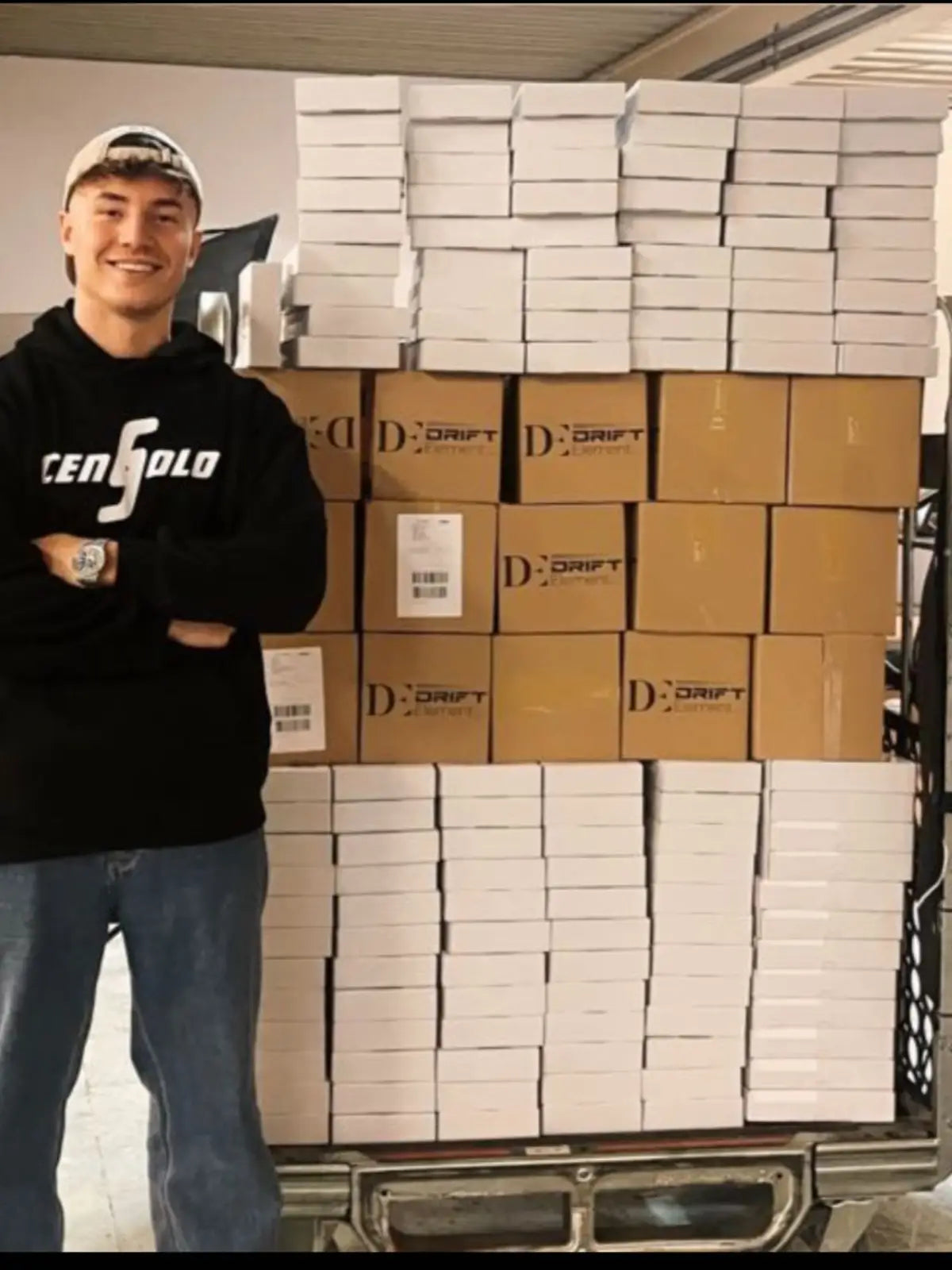 The image features a young man smiling and posing in front of a large stack of boxed products. On his wrist is a stylish watch, likely one of the rim-designed watches by DriftElement, indicative of the company's innovative product design. The boxes are labeled with the DriftElement logo, suggesting a successful batch of orders ready for shipment from this young and dynamic German startup.